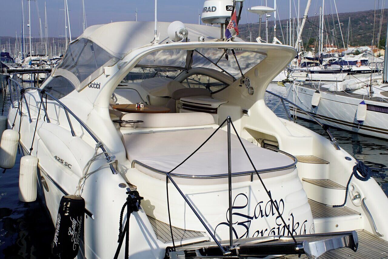Un yate blanco con la inscripción 'Radyo Marina' en la proa, amarrado en un puerto deportivo con otros barcos.