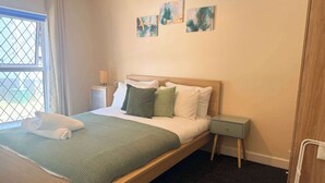 Ein ordentlich bezogenes Bett mit grüner Decke und Kissen, ein Nachttisch mit Lampe und Kunstwerk an der Wand.