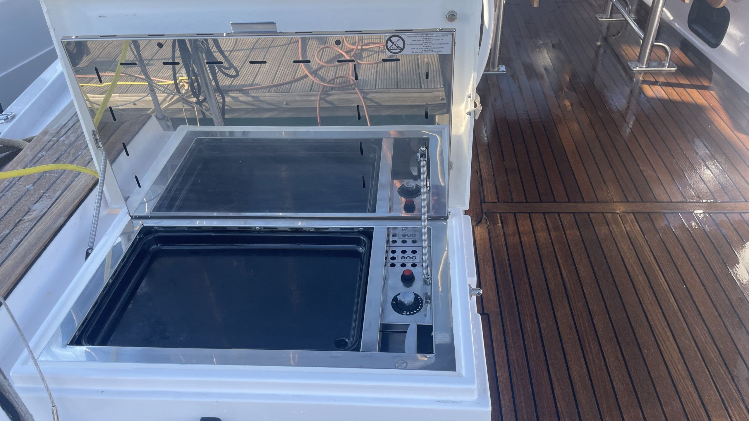 An open, metallic, rectangular appliance with control panels and hoses, situated on a wooden deck.