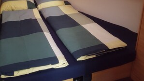 A compact bed with a striped bedspread, a small window above, and a wooden bench to the right.