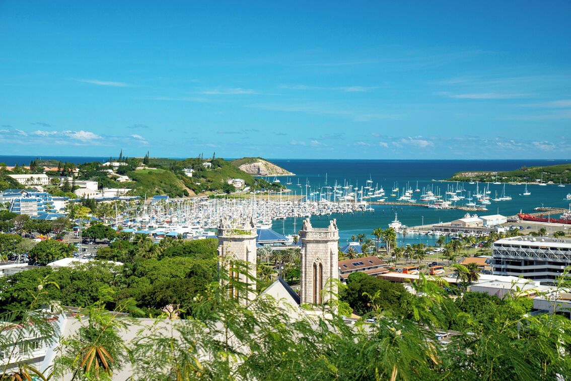 Un port de plaisance où sont amarrés de nombreux bateaux, une église à deux tours et une ville côtière avec des bâtiments et de la verdure.