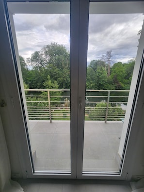 Une vue à travers une porte vitrée donnant sur un balcon extérieur, avec une rambarde en métal, de la verdure et un ciel nuageux.
