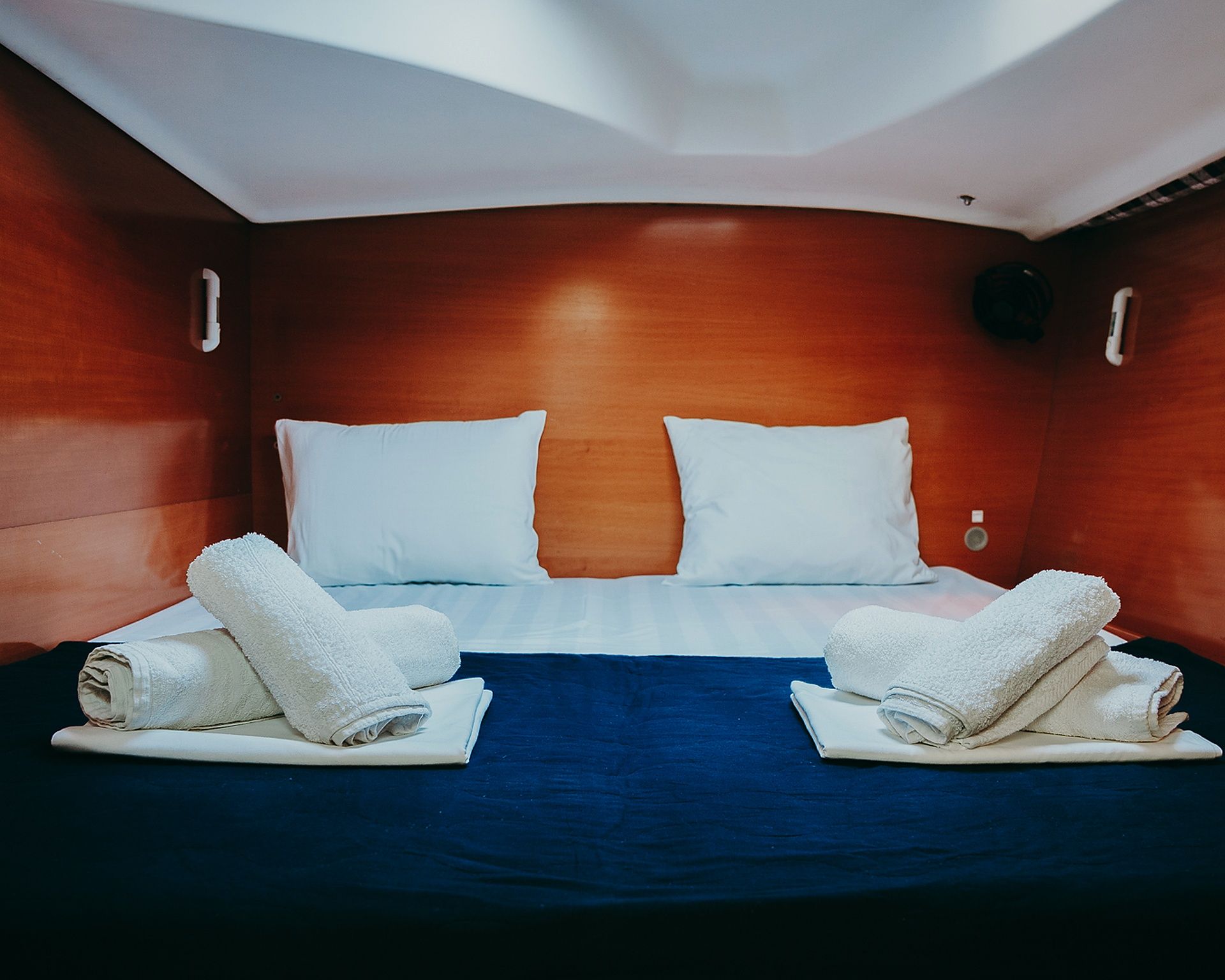 Habitación de hotel con cabecera de madera, dos almohadas blancas, colcha azul y dos toallas enrolladas sobre la cama.