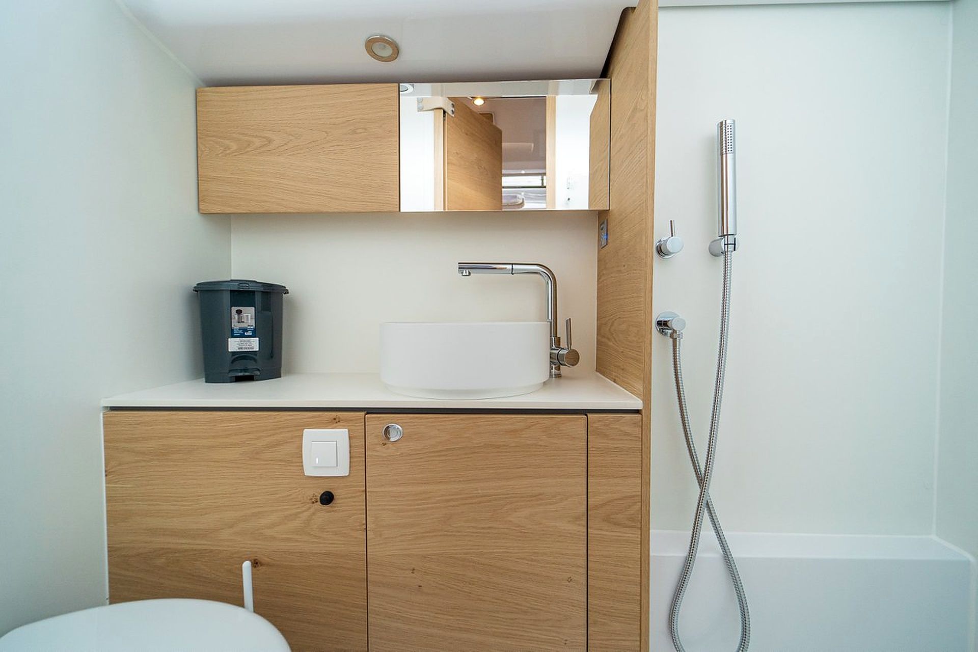 A bathroom with a wooden vanity, a white sink, a mirror, a shower, and a trash bin.
