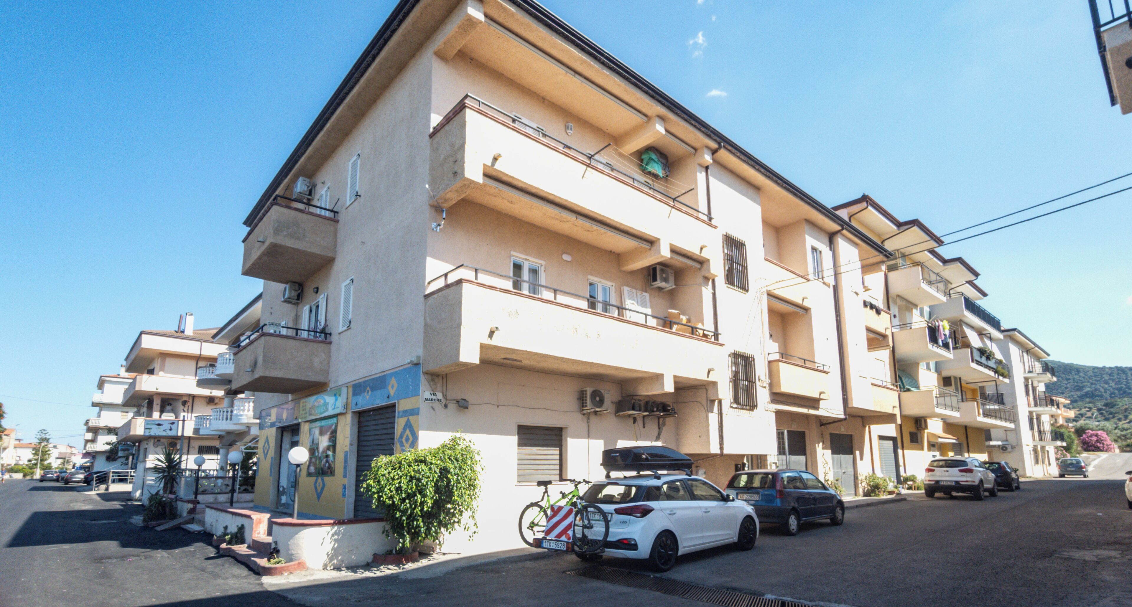 Un immeuble résidentiel à plusieurs étages, doté de balcons, avec des voitures garées et un ciel dégagé.