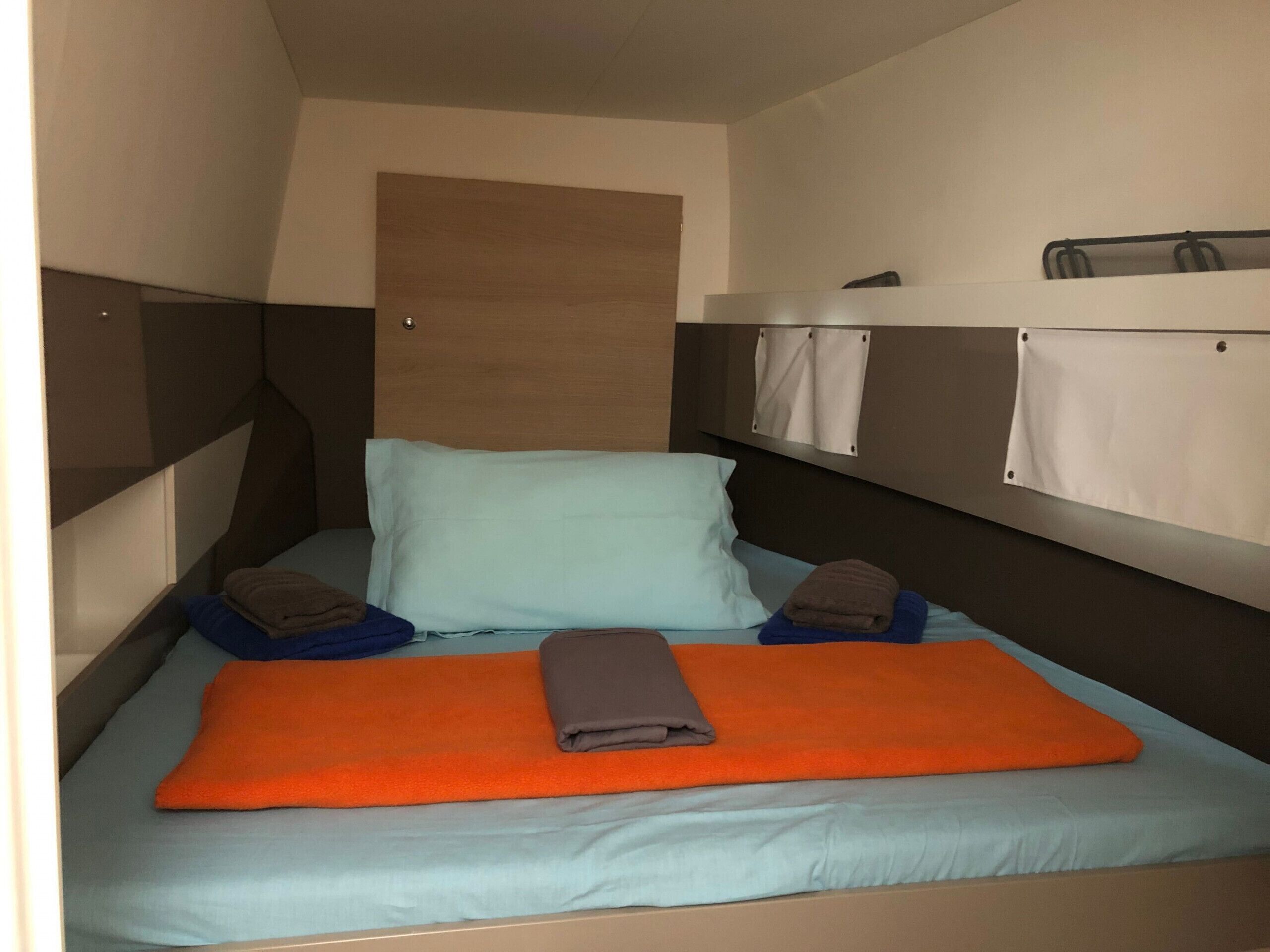 Een eenpersoonsbed met een blauwe matras, twee kussens, een oranje deken en een bruine handdoek. Er zijn twee opbergvakken voorzien van witte gordijnen.