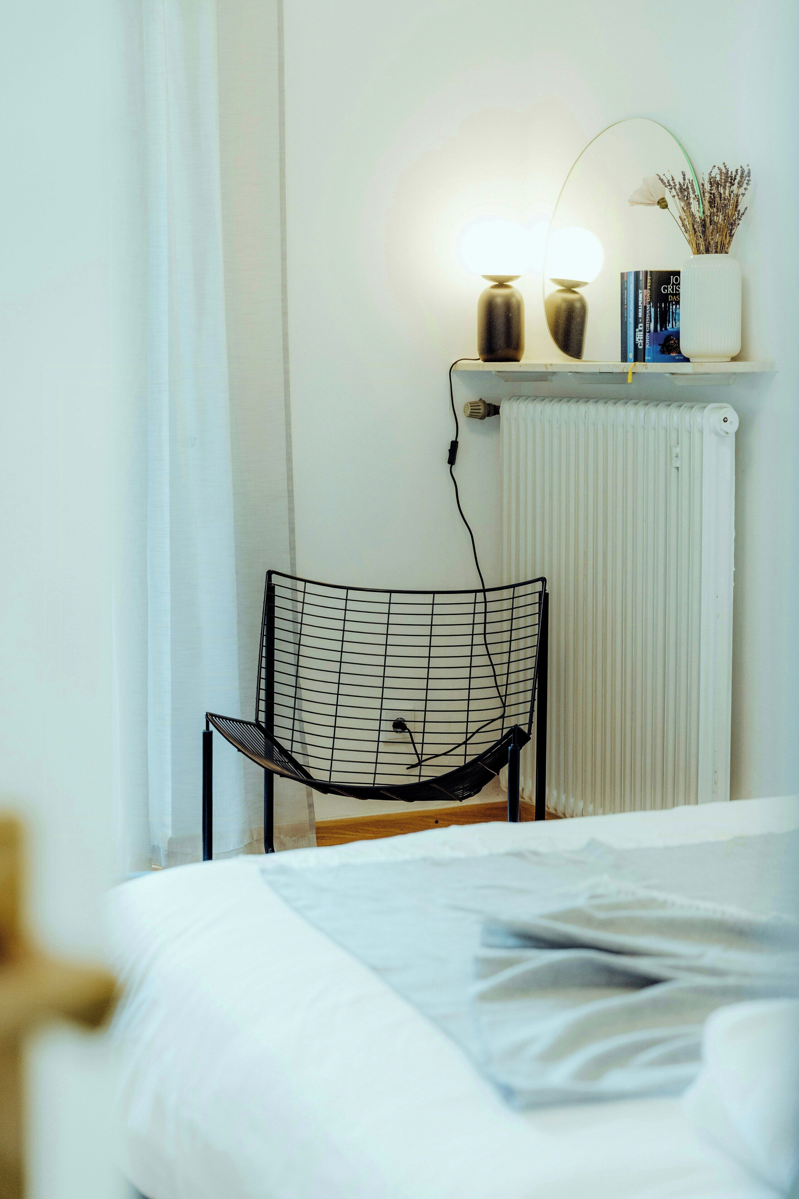 A modern room with a bed, a black wireframe chair, a white radiator, and a shelf with books and a plant.
