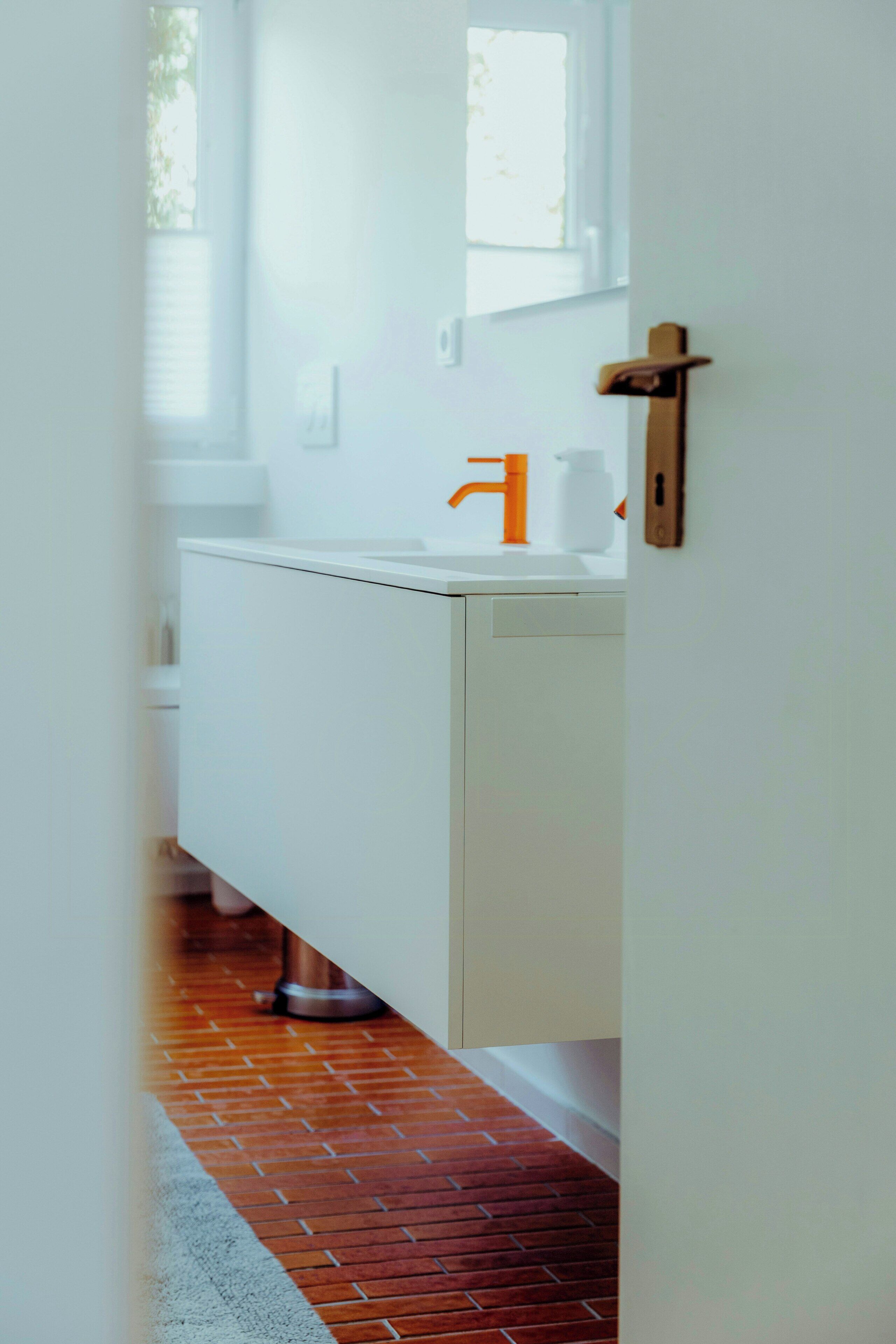 A bathroom with a white sink, an orange faucet, and a metal floor vent.