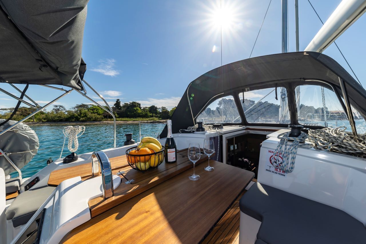 Een dek van een boot met een houten tafel, een fruitschaal, wijnglazen en een fles.