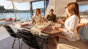 Uma área de refeições em um barco, com uma mesa posta para quatro pessoas, rodeada de cadeiras, e vista para a água e colinas ao fundo.