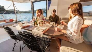 A dining area on a boat with a table set for four, surrounded by chairs, with a view of the water and hills in the background.