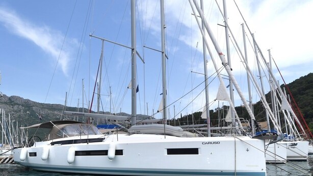 Eine weiße Yacht namens 'CARUSO' liegt in einer Marina vor Anker, gesäumt von mehreren Masten und Flaggen, umgeben von anderen Booten und einer gebirgigen Landschaft.