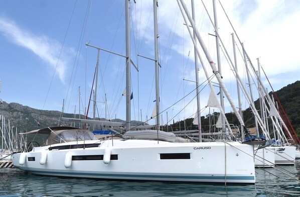 Un yate blanco llamado 'CARUSO' amarrado en un puerto deportivo con numerosas antenas y banderas, rodeado de otras embarcaciones y un paisaje montañoso.