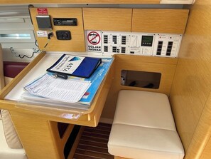 A compact cabin with a wooden table holding documents, a blue folder, and a map. A small white seat is visible, and a control panel with various switches and indicators is mounted on the wall. - Sun-Kissed Sailing Adventure in Zadar, Croatia (Bibinje)