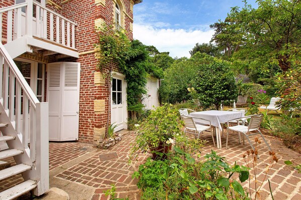A brick house with a white balcony, a garden with a table and chairs, and a brick pathway.