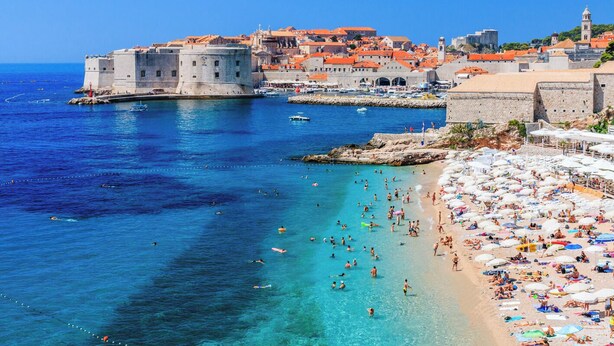 A coastal city with a historic fort, clear blue waters, and a crowded beach.