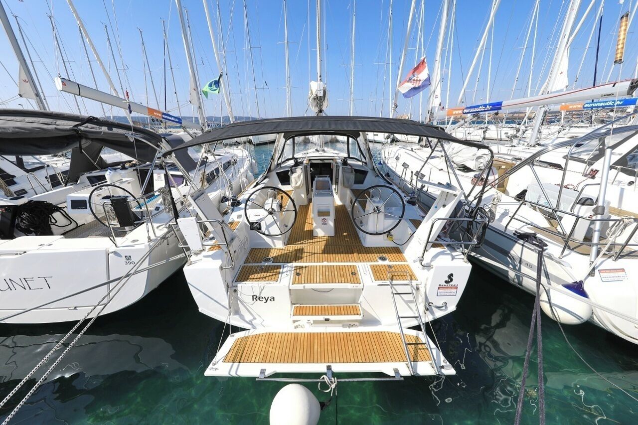 A marina with multiple sailboats docked, featuring a wooden deck, a steering wheel, and the name 'Reya' on the bow.