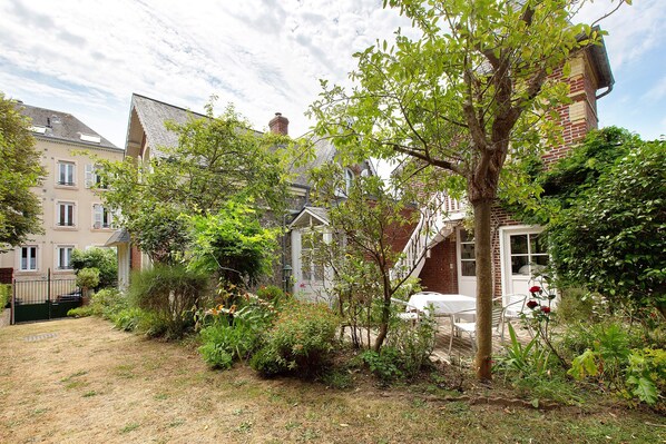A residential garden with a lawn, various plants, a tree, and a white table with chairs.