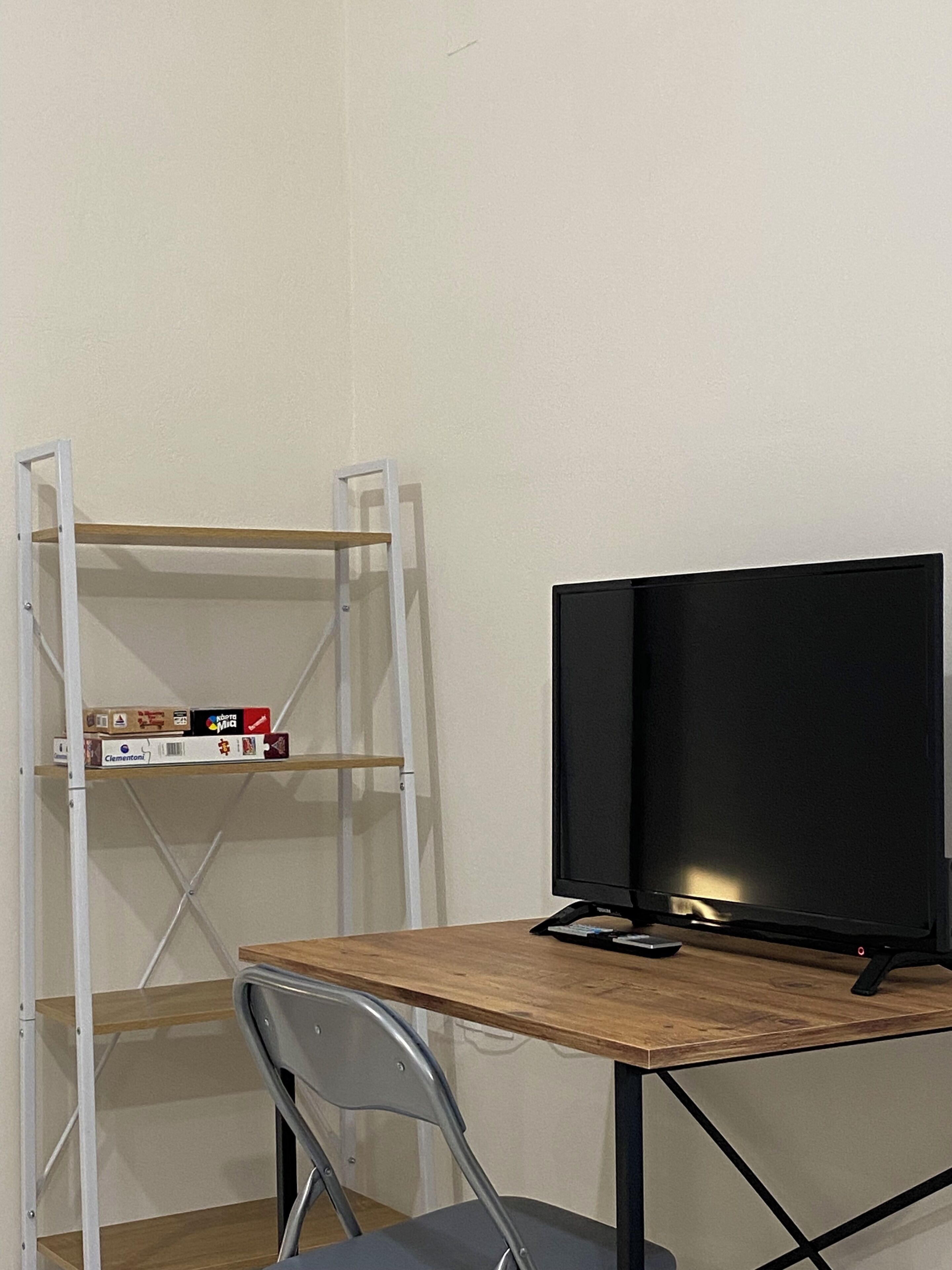 Une pièce comprenant un bureau, une chaise, un téléviseur et une étagère blanche sur laquelle sont posés quelques objets.