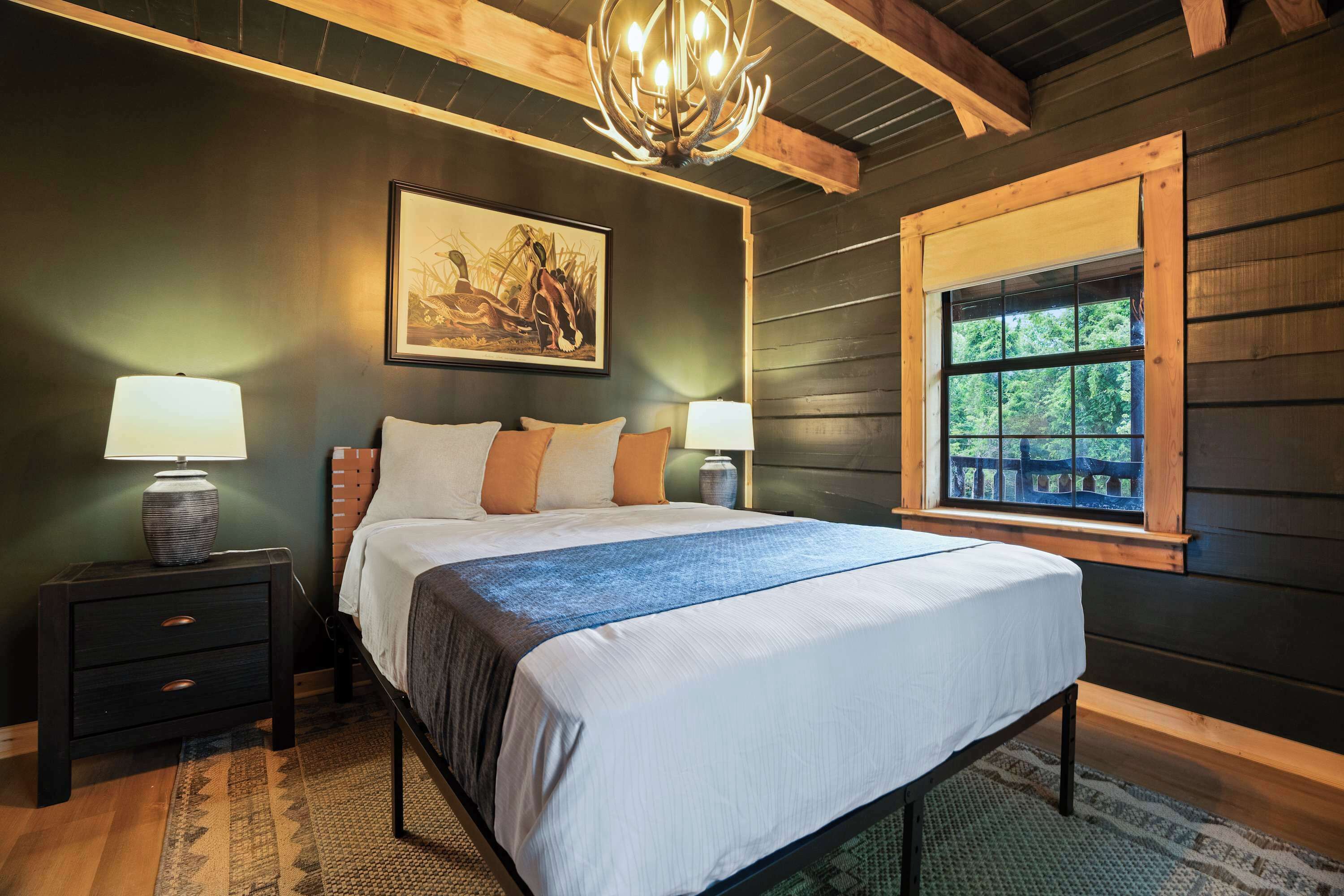 A bedroom with a bed, two bedside tables, a chandelier, a window with a view, and a painting on the wall.