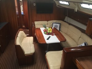 A cozy interior with a cushioned seating area, a wooden table with fruits and a bottle, and a mounted television. - Sun-Kissed Sailing Adventure in the Ionian Islands (Nidri)