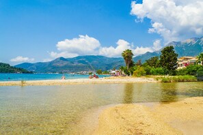 Ein Strand mit klarem Wasser, sandigem Ufer und Bergen im Hintergrund.