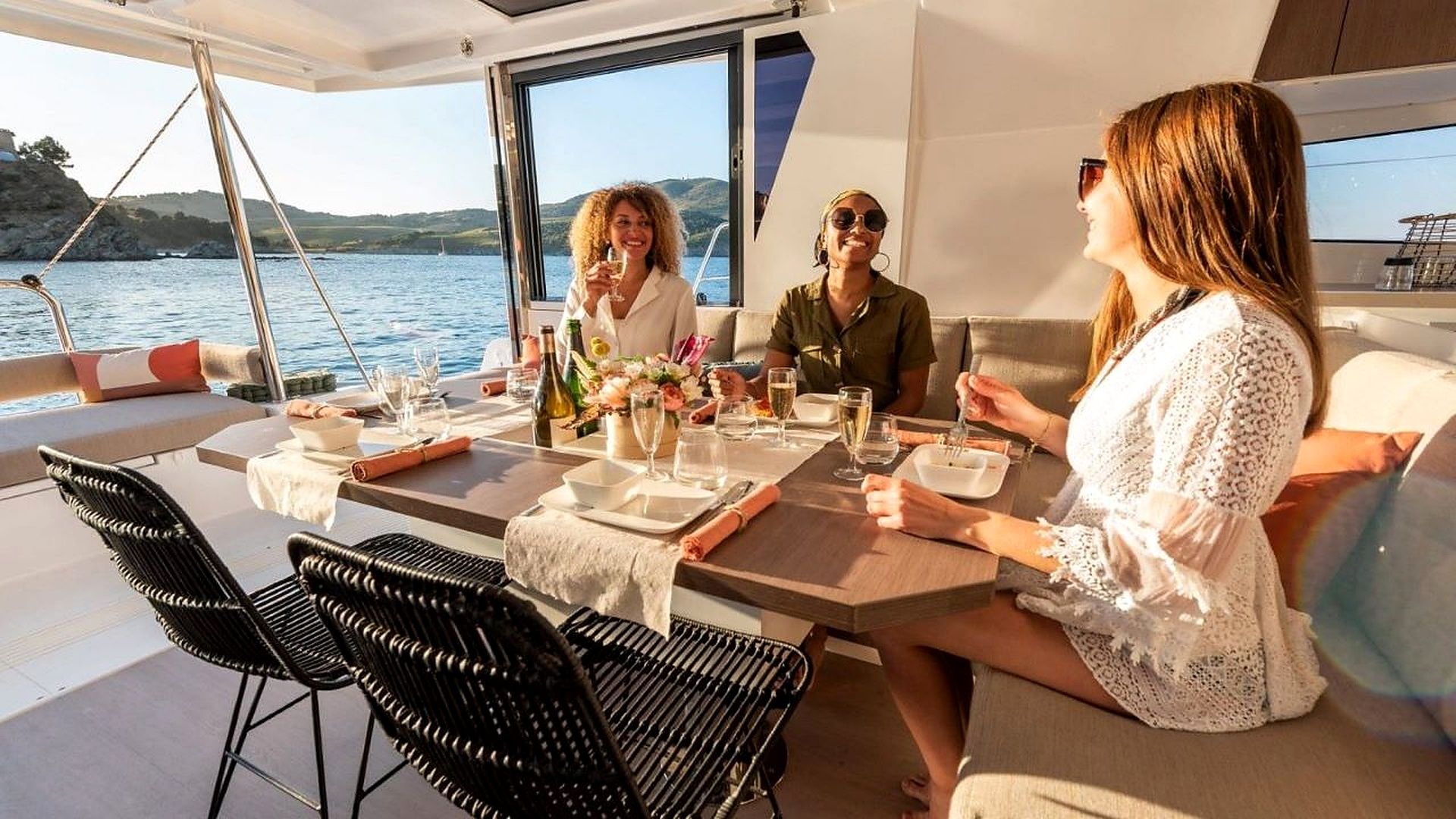 Un coin repas sur un bateau, avec une table dressée pour quatre personnes, entourée de chaises, et une vue sur l’eau et les collines en arrière-plan.