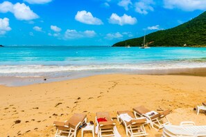 A beach with clear turquoise water, a sandy shore, and several lounge chairs arranged for sunbathing.