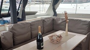 A cabin with a cushioned seating area, a table with a bottle and a decorative basket, and a view of the marina through a window. - Sun-kissed Sailing Adventure in Split-Dalmatia, Croatia (Seget Donji)