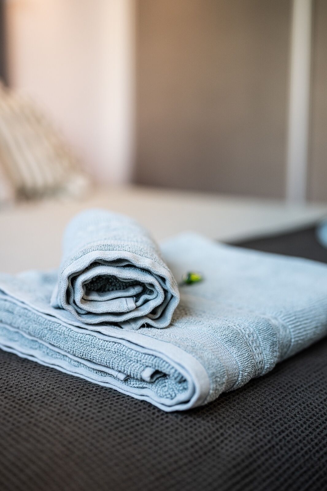 A folded towel on a textured surface, possibly a bed or couch, with a blurred background.