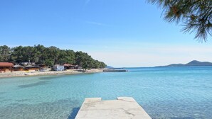 A clear waterfront with a concrete dock, a beach with buildings, and a forested hill in the background. - Sun-Kissed Sailing Adventure in Zadar, Croatia (Biograd na moru)