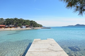 A clear waterfront with a concrete dock, a beach with buildings, and a forested hill in the background.