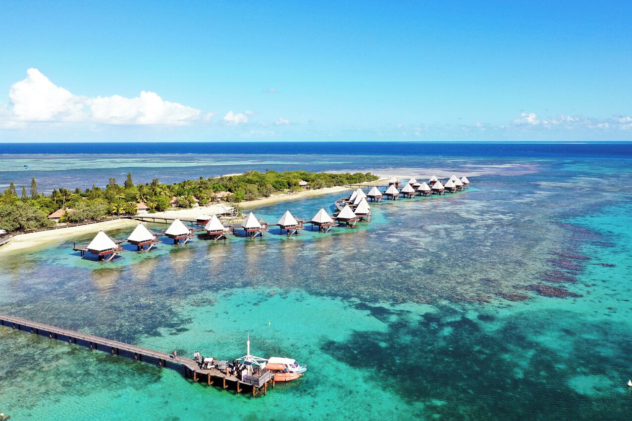 Vista aérea de bungalows sobre el agua conectados por un muelle, rodeados de aguas turquesas cristalinas y una costa lejana.