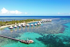 Vista aérea de bungalows sobre el agua conectados por un muelle, rodeados de aguas turquesas cristalinas y una costa lejana.