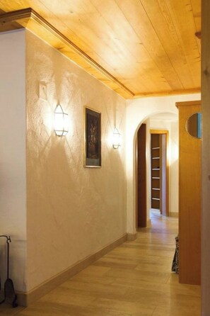 Een hal met houten plafond en wanden, twee wandlampen, een ingelijst kunstwerk en een deur naar een andere kamer.