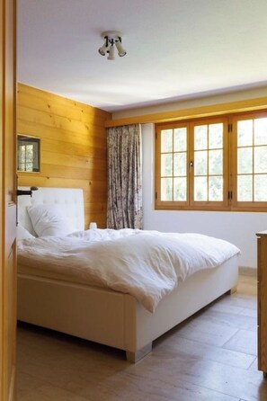 A bedroom with a wooden floor, a bed with white bedding, a window with curtains, and a ceiling light fixture. - Graebi Appartement/Fewo, Dusche und Bad, WC 8-Bettwohnung by Interhome (Gstaad)