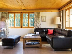 Een woonkamer met een houten plafond, een zwarte leren bank, een houten salontafel en een raam met gordijnen.