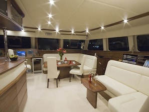 A yacht interior with a dining area, a sofa, a small table with a bottle and glass, and a control panel.