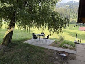 An outdoor seating area with a table and chairs, a fire pit, and a view of a green landscape with trees and a building in the distance. - Hivernants Le Premier Appartement/Fewo, Dusche und Bad, WC 4-Bettwohnung by Interhome (Gstaad)