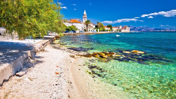 Uma paisagem costeira com água cristalina, uma praia e edifícios ao fundo.