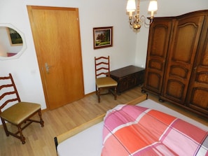 Ein Zimmer mit Holzschrank, Holzstuhl, Bett mit gestreifter Bettdecke, kleinem Tisch und einem an der Wand hängenden Bild.