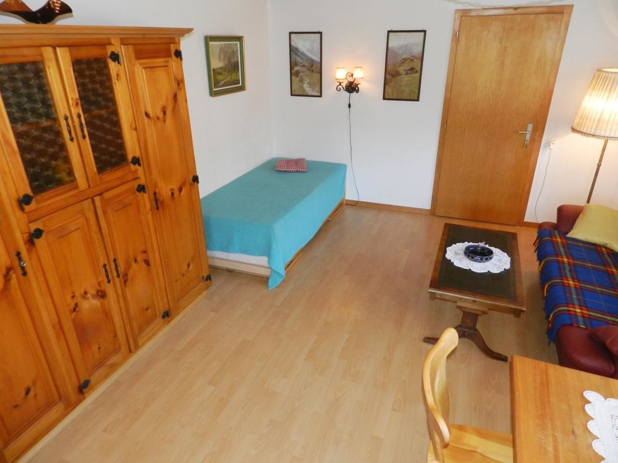 Um quarto com uma cama, um armário de madeira, uma mesa com um jogo americano de renda e um sofá com um cobertor xadrez.