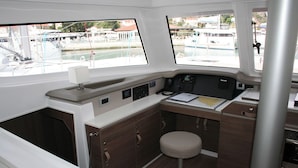 Eine kleine Kombüse mit einer zentralen Bedienkonsole, einem kleinen Hocker und Blick auf den Hafen durch Fenster.