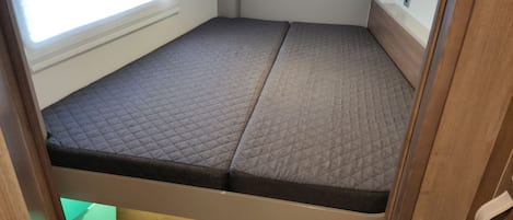 A compact bed with a quilted mattress, a window with blinds, a white wall, and a wooden cabinet.