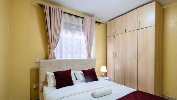 A bedroom with a bed, pillows, a nightstand, a lamp, a window with curtains, and a wardrobe. - Cozy, secure home stay with modern comforts and a convenient location. (Kisii County)