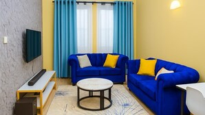 Ein Wohnzimmer mit einem blauen Sofa, einem runden Couchtisch, einem an der Wand befestigten Fernseher und einem Fenster mit blauen Vorhängen.