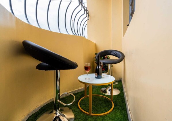 Una habitación pequeña con una pared amarilla, una silla negra, una mesa redonda con una botella y un vaso, y una ventana con barandilla de metal.