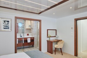 Une chambre d’hôtel moderne dotée d’une salle de bain entièrement vitrée, d’une coiffeuse en bois, d’un lit et d’un grand miroir.