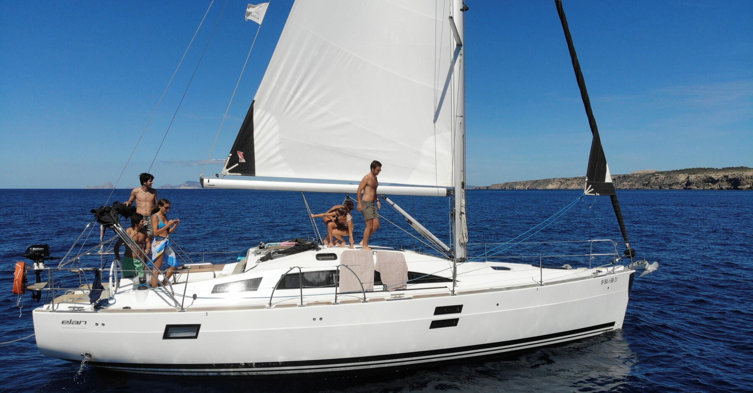 Een zeilboot met een witte romp en zwarte accenten, uitgerust met een motor en zeilen, afgemeerd in een jachthaven met rotsachtig terrein op de achtergrond.