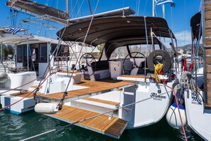 Ein Yachthafen mit einer weißen Yacht namens 'Haman', die an einem Steg liegt. Man sieht ein überdachtes Deck, Holzboden und verschiedene Seile und Ausrüstung.